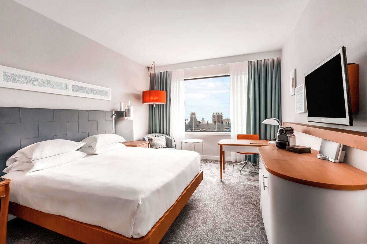 A hotel room with a bed, a desk, a chair, a TV, and a window with a city view.