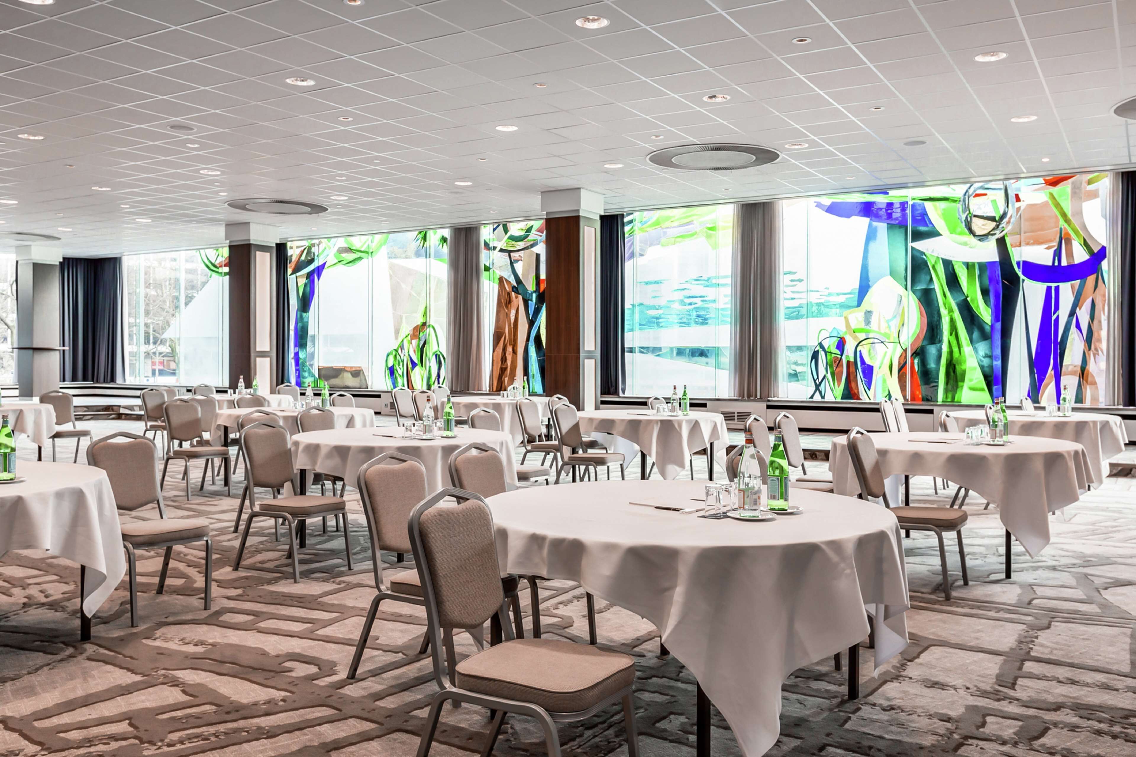 A spacious banquet hall with round tables and chairs, large windows with abstract art, and a patterned carpet.
