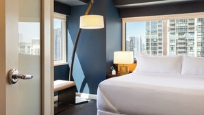 Ein Hotelzimmer mit einem großen Bett, einem Schreibtisch mit Lampe, einer Bank und Blick auf die Skyline durch große Fenster.