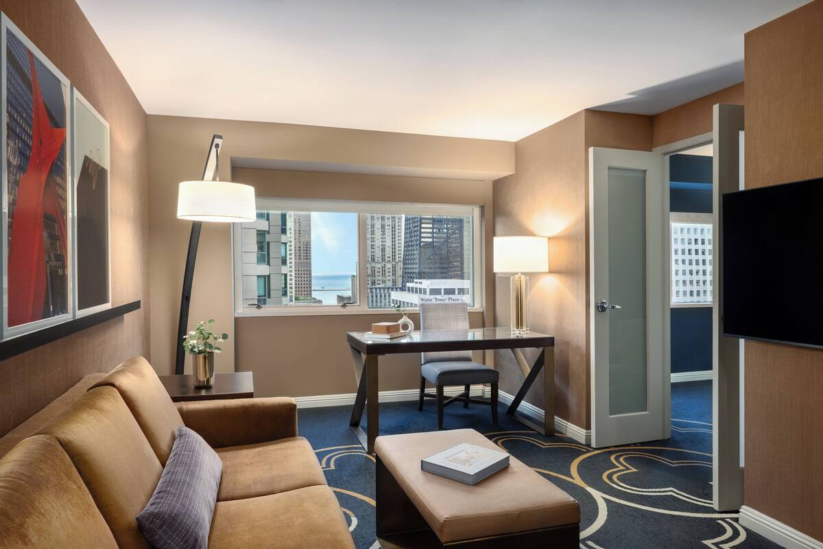 A hotel room with a sofa, a desk, a chair, a TV, and a window with a city view.