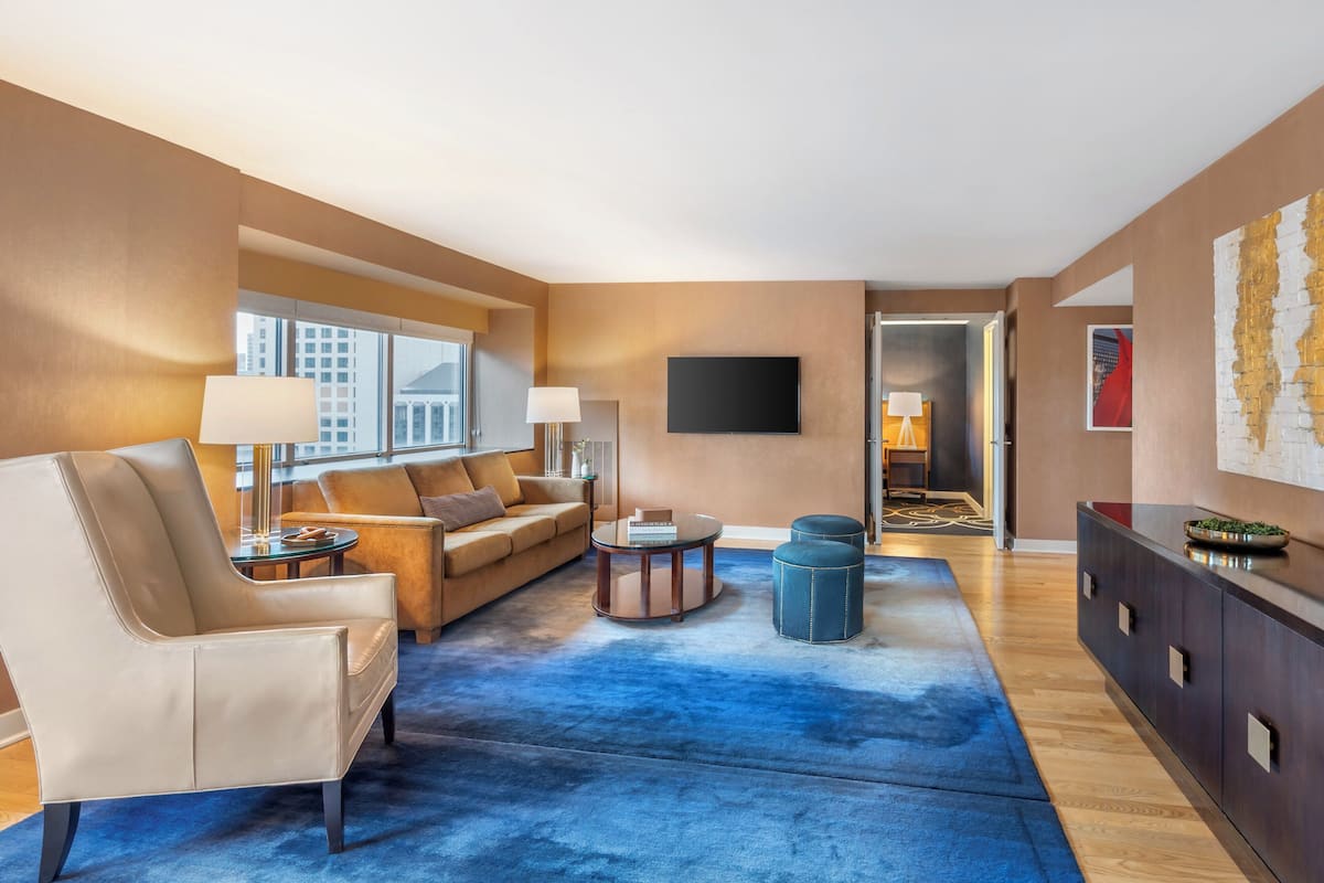 A modern living room with a sofa, armchair, and coffee table, a blue carpet, and a TV mounted on the wall.