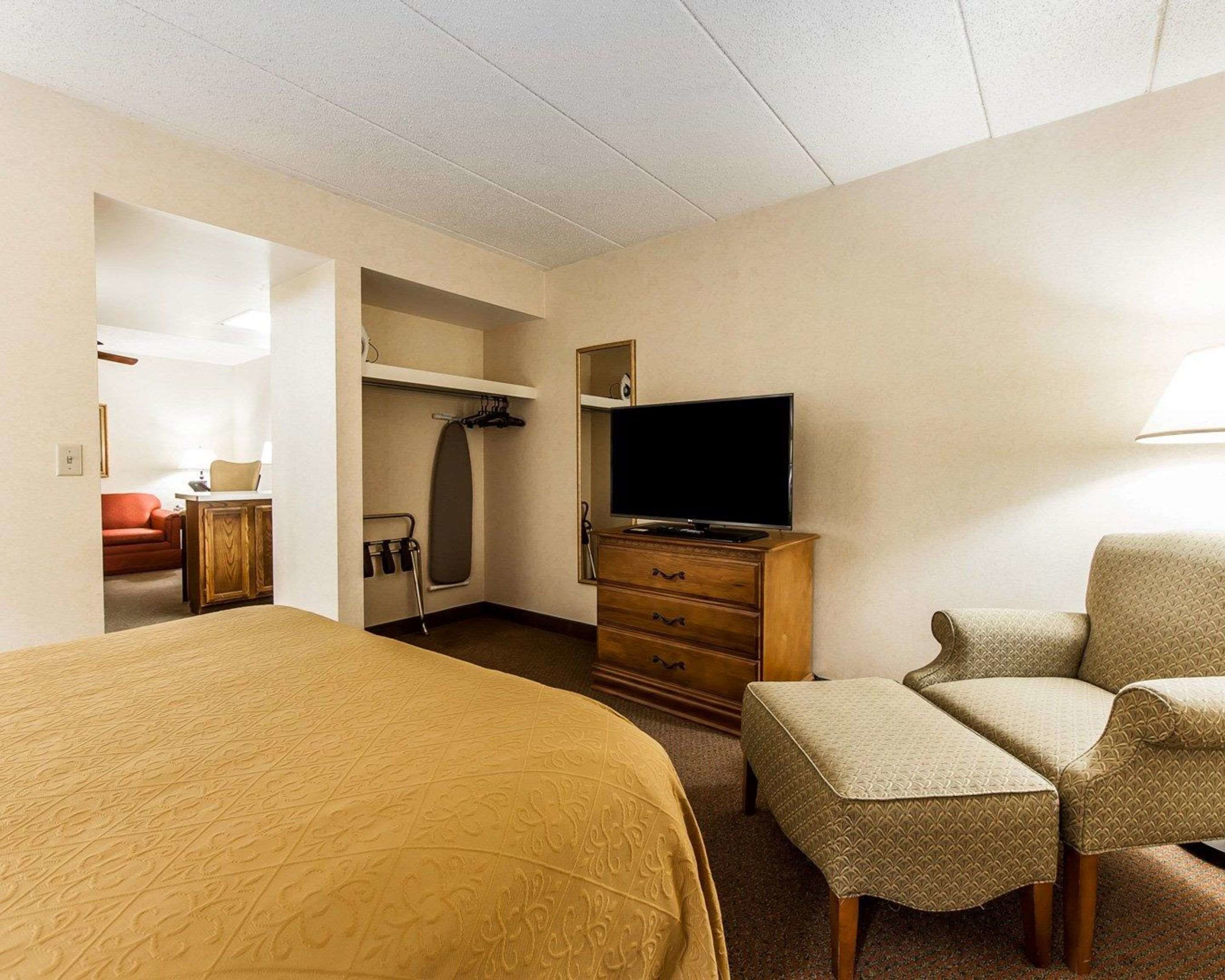 A hotel room with a large bed, a television, a desk with a chair, a wardrobe, and a seating area with a chair and ottoman.
