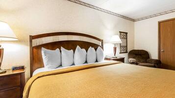 A hotel room with a large bed, a wooden headboard, a nightstand with a lamp, a chair, and a door.