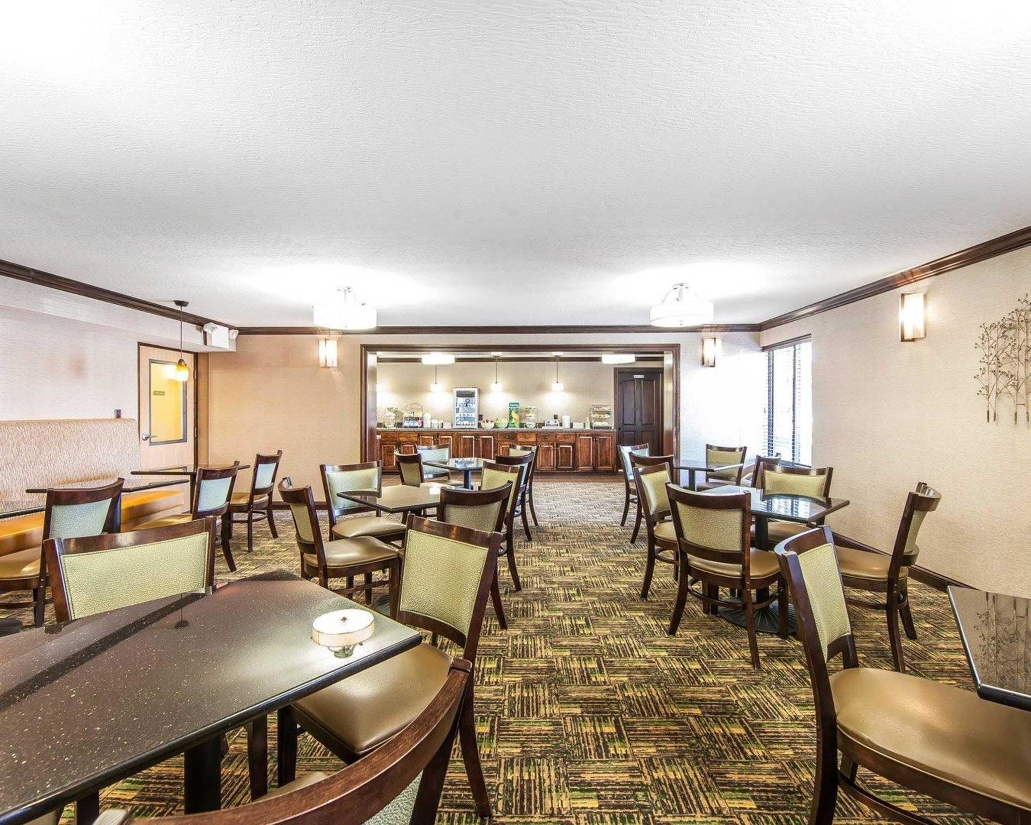 A hotel dining area with multiple tables and chairs, a kitchen in the background, and a patterned carpet.