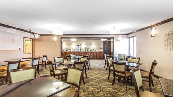 Un comedor de hotel con varias mesas y sillas, una cocina al fondo y una alfombra estampada.