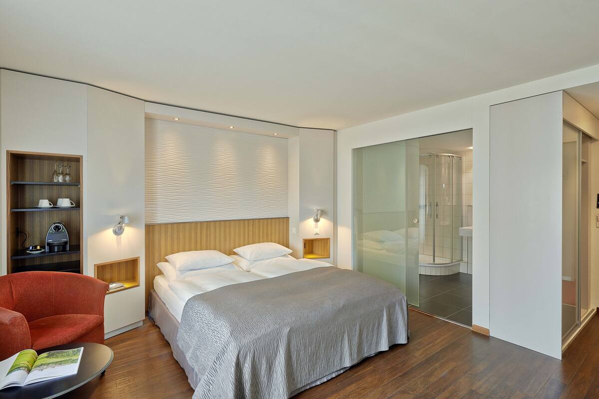 Una habitación de hotel moderna con una cama grande, un sillón rojo, una mesita con una revista, un estante de madera con utensilios de cocina y un box de ducha de vidrio.