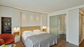 A modern hotel room with a large bed, a red armchair, a small table with a magazine, a wooden shelf with kitchen items, and a glass shower enclosure.