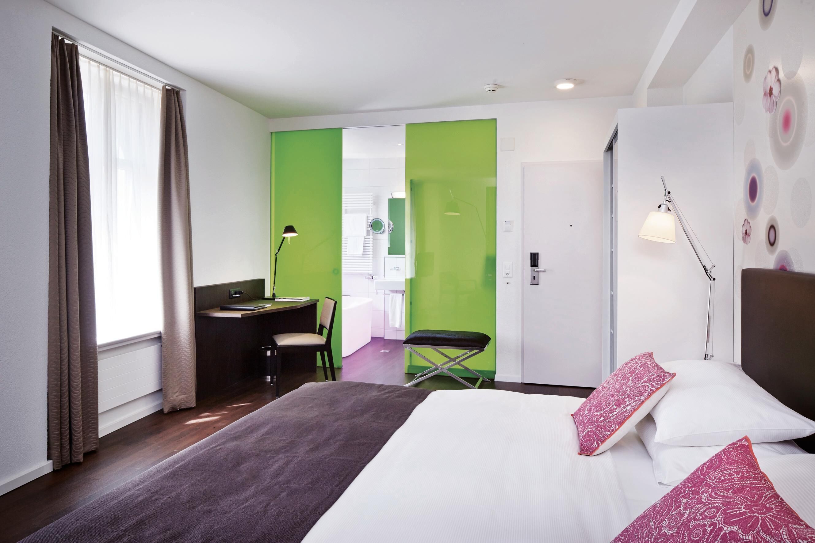 Habitación de hotel con cama, escritorio, silla, banco, lámpara y vista al baño.