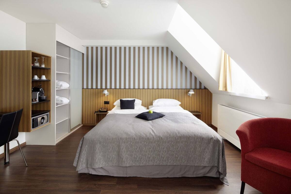 Una habitación de hotel moderna con techo inclinado, una cama con colcha gris estampada, un sillón rojo y un armario de madera con estantes.