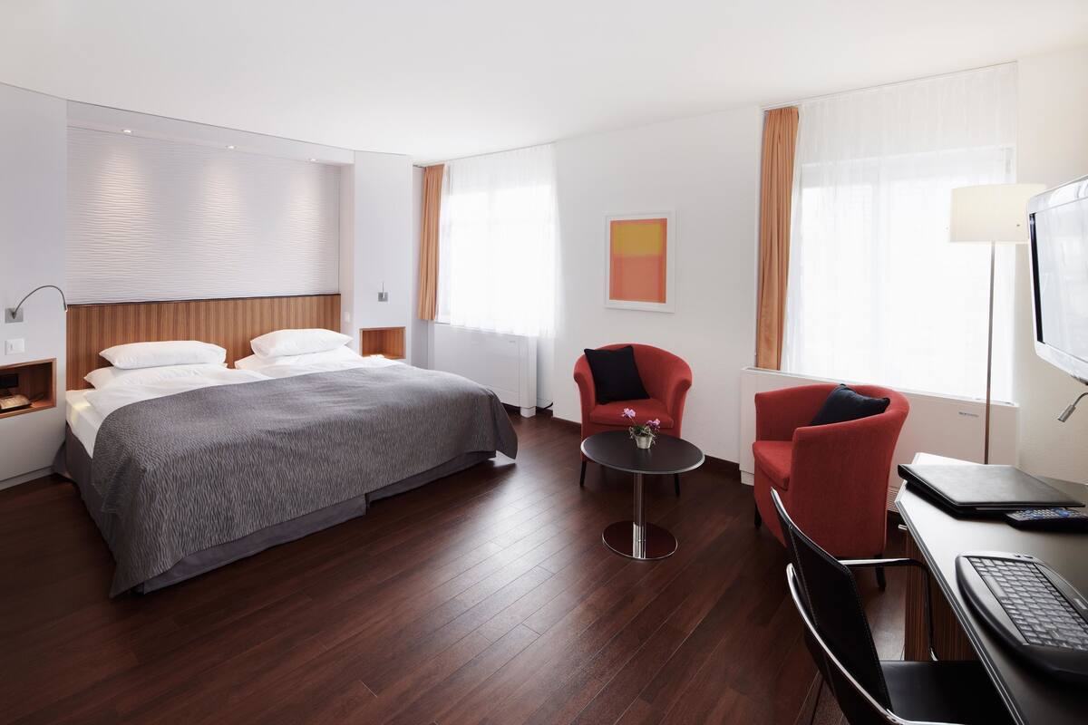 Habitación de hotel con cama, escritorio con una computadora portátil, silla y una mesita con un florero.