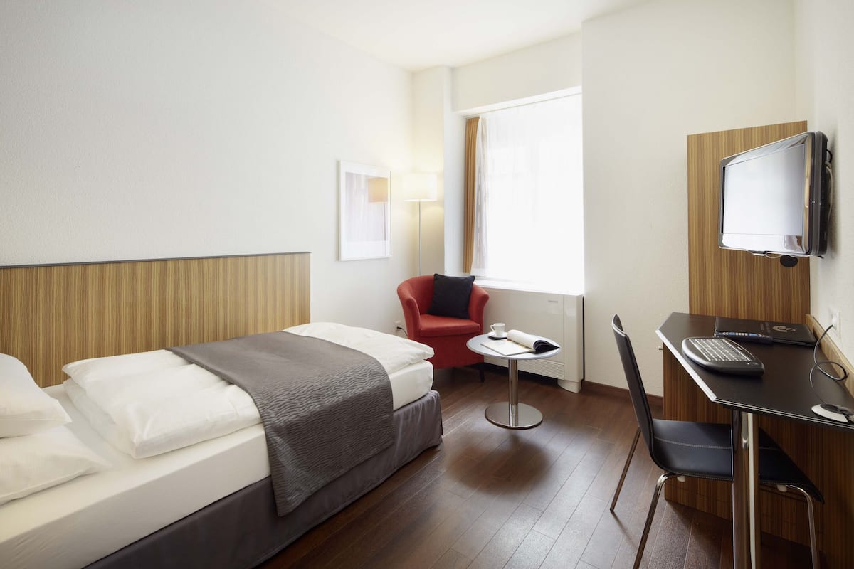 Habitación de hotel con cama, escritorio con computadora portátil, silla, televisor montado en la pared y ventana con cortinas.