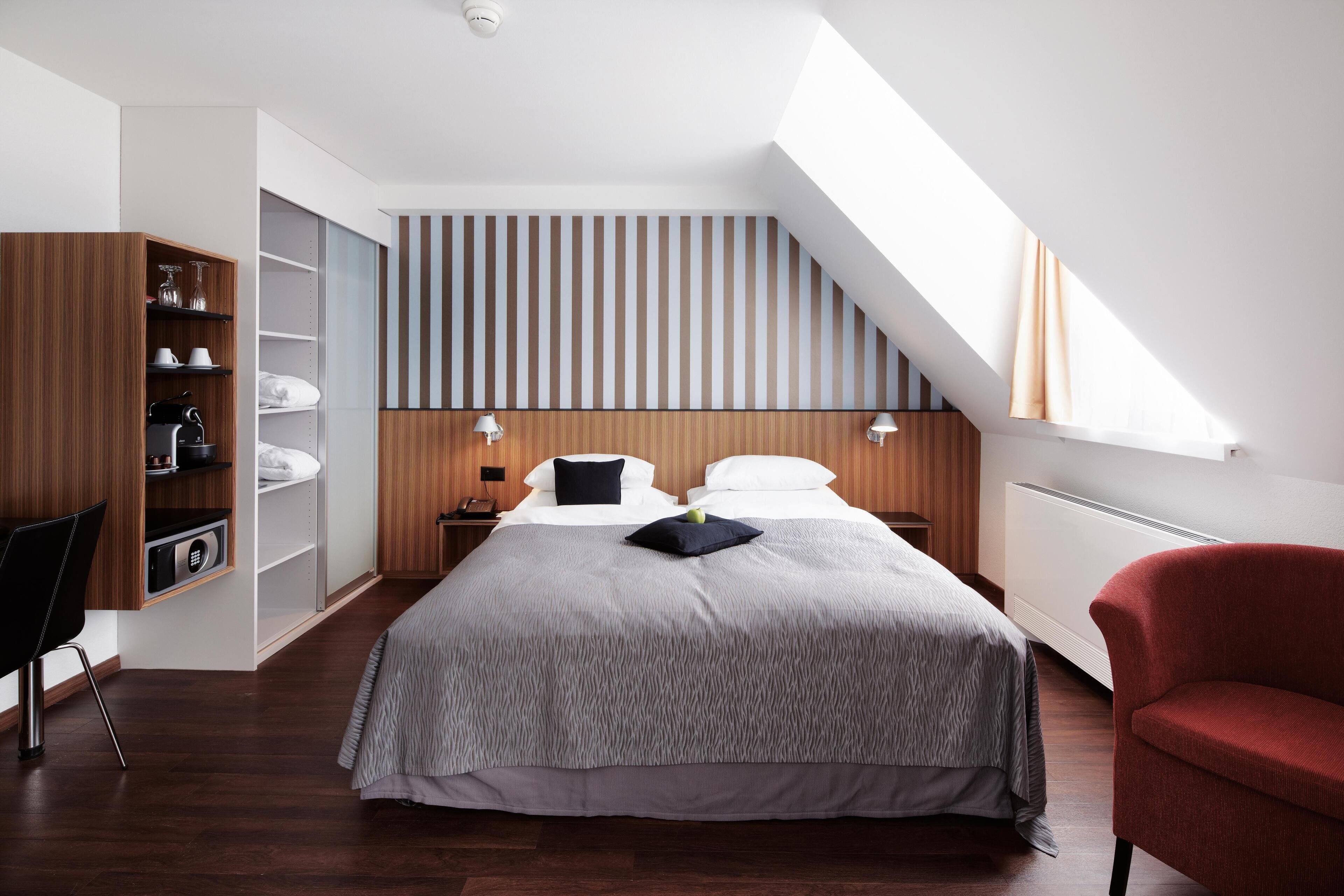 A modern bedroom with a sloped ceiling, a large bed with a grey patterned bedspread, a red armchair, and built-in wooden shelving with various items.