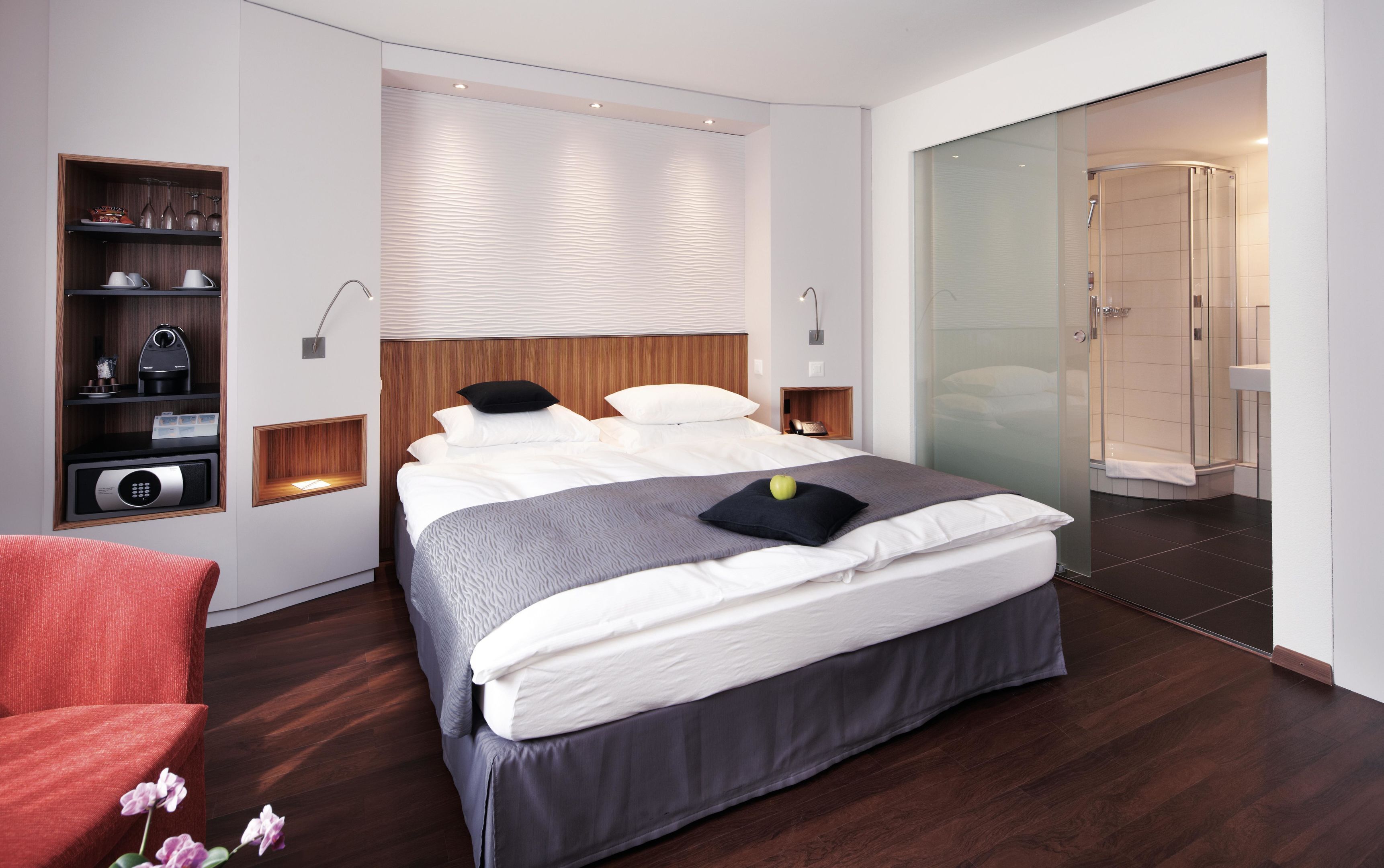 A modern bedroom with a large bed, bedside tables, a built-in shelf with kitchen appliances, and a glass shower enclosure.