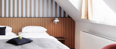 Una habitación de hotel moderna con techo inclinado, cama con edredón gris texturizado, cabecera de madera, sillón rojo y lámpara fijada a la pared.