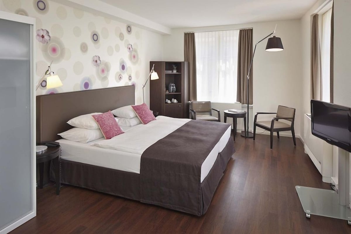 Habitación de hotel con cama, mesitas de noche, escritorio, silla, televisión, estantería y ventana con cortinas.