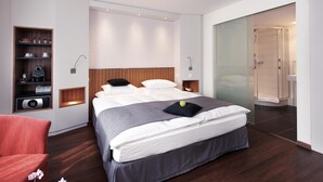 A modern bedroom with a large bed, a built-in shelf with kitchen appliances, a glass shower enclosure, and a red chair.