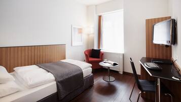 A hotel room with a bed, a desk with a computer, a chair, a TV mounted on the wall, and a window with curtains.