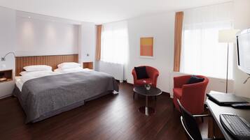 A modern hotel room with a large bed, a desk with a laptop, a red armchair, and a small table with a vase.