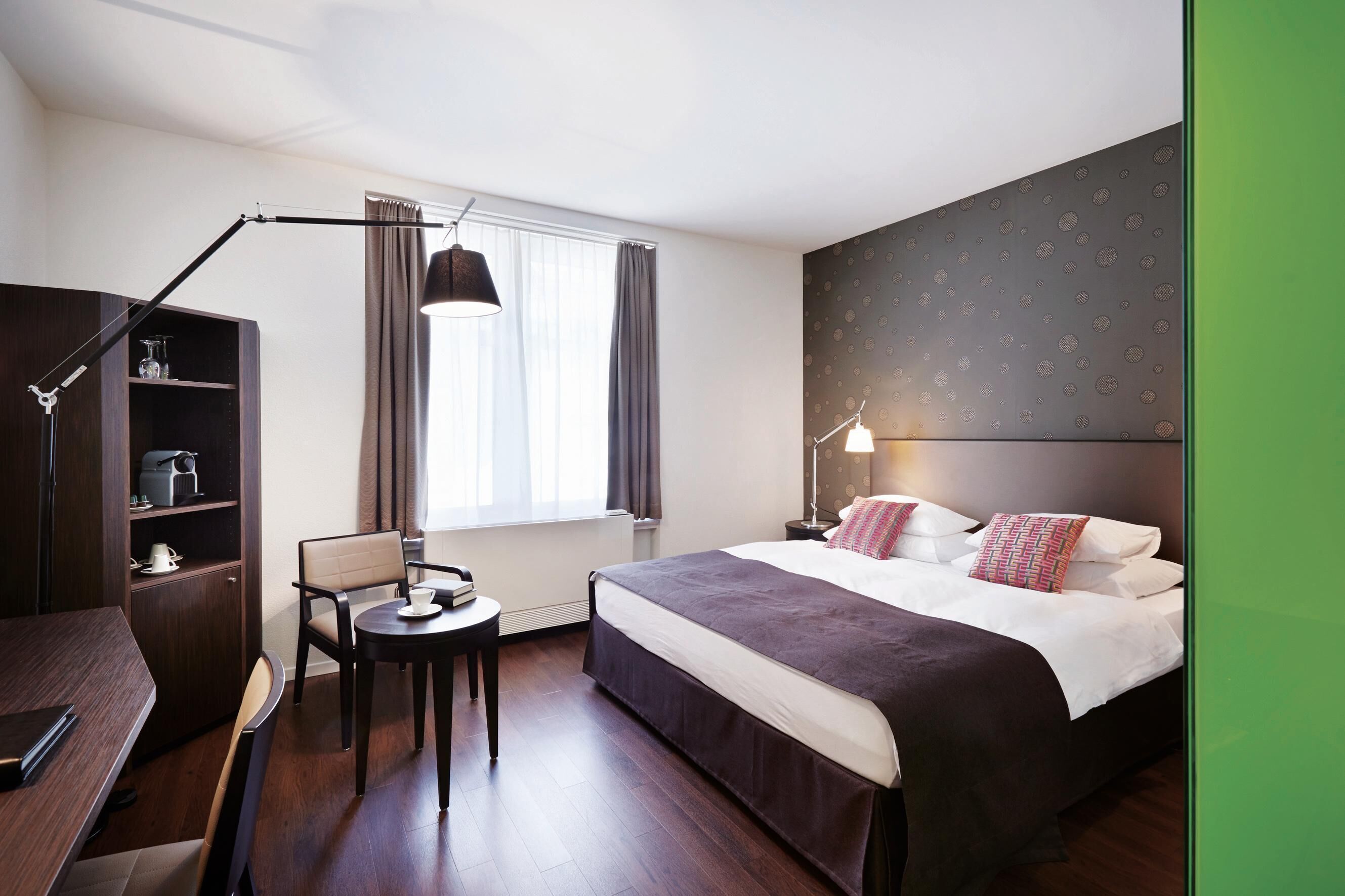 Una camera d'albergo con un letto, comodino, lampada, sedia, scrivania e finestra con tende.