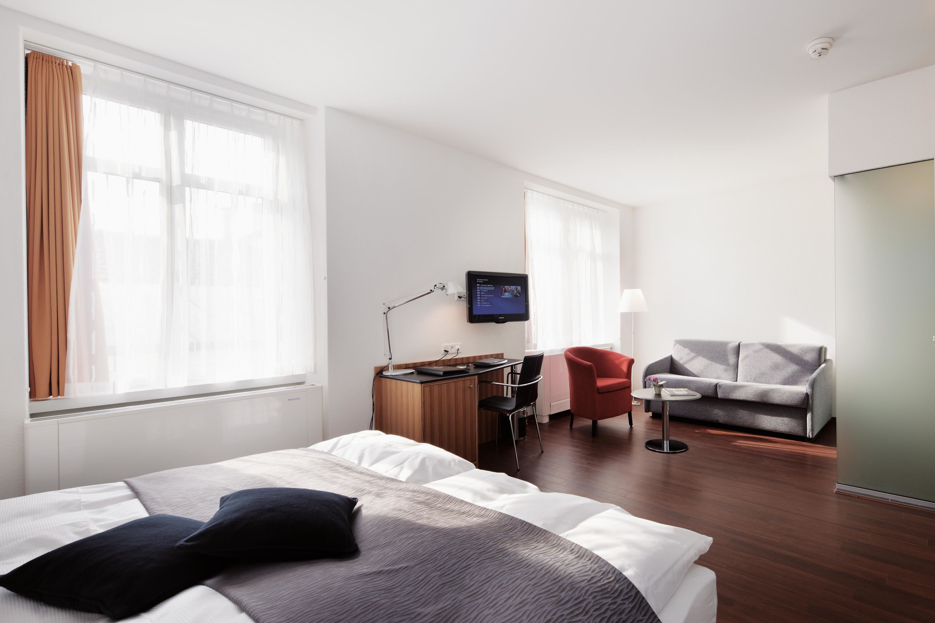 A hotel room with a bed, a desk with a computer, a TV, a red chair, a grey sofa, and large windows.