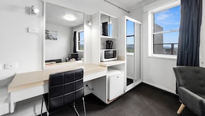 Habitación de hotel moderna con escritorio blanco, silla negra, microondas, refrigerador pequeño y ventana grande con vista a la ciudad.