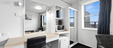A modern hotel room with a white desk, a black chair, a microwave, a small refrigerator, and a large window with a view of the city.