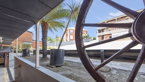 Vista a través de una ventana con una gran rueda de madera, una palmera y un edificio de ladrillo al fondo.