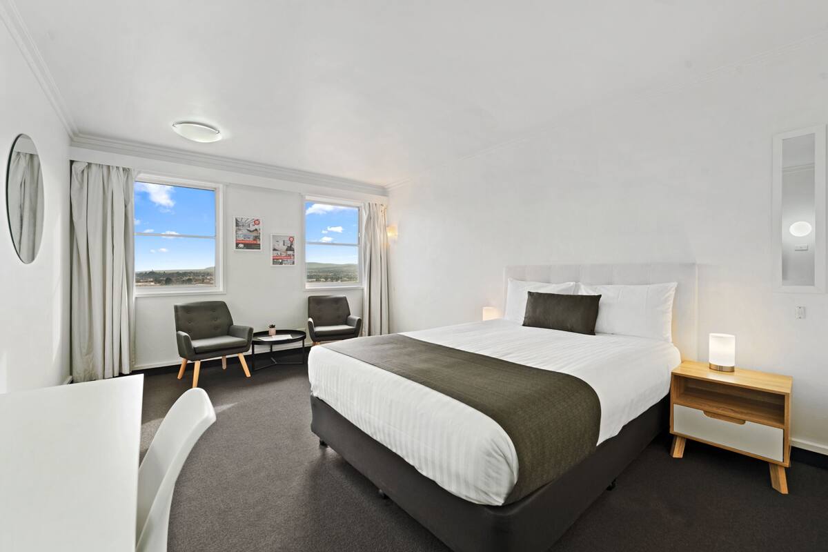 A hotel room with a large bed, two armchairs, a desk, and a nightstand with a lamp.