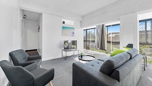 Una sala moderna con un sofá gris, dos sillones, una mesita de centro y un televisor. Hay un ventanal con vistas al exterior.