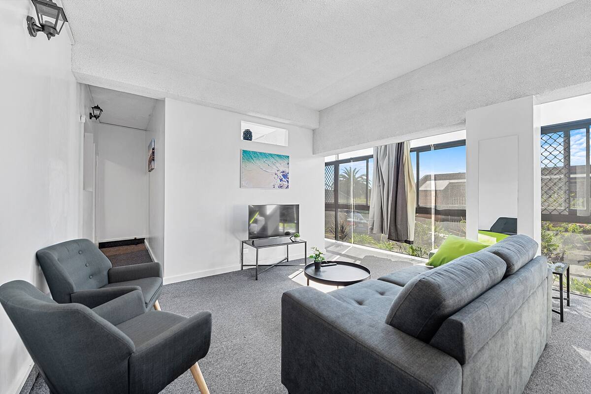A modern living room with a grey sofa, two armchairs, a coffee table, and a TV. There is a large window with a view of the outdoors.