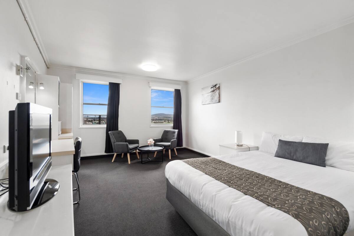 A hotel room with a large bed, two windows with curtains, a desk with a computer, two chairs, and a TV.