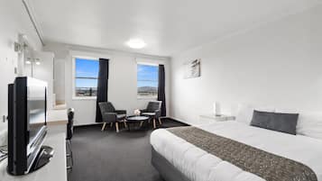 A hotel room with a large bed, two windows with curtains, a desk with a computer, two chairs, and a TV.