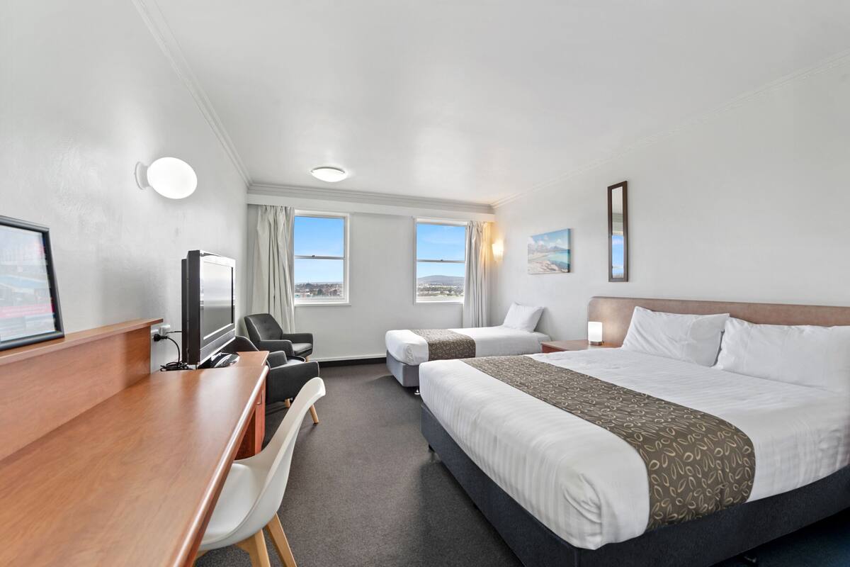 A hotel room with two beds, a desk, a chair, a TV, and large windows with a view.