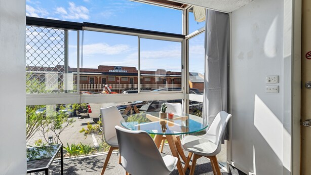 Una habitación con mesa de vidrio y sillas blancas, un ventanal con vista a edificios y un estacionamiento.