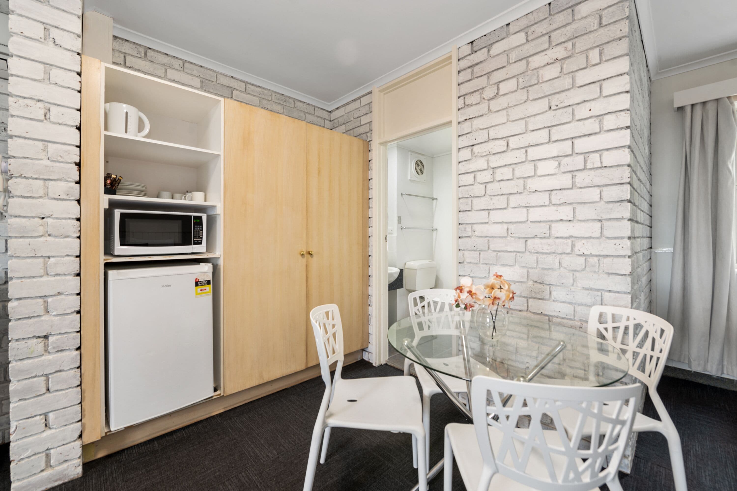 Una habitación compacta con kitchenette, una zona de comedor con una mesa de vidrio y sillas blancas, y una pared con diseño de ladrillo.