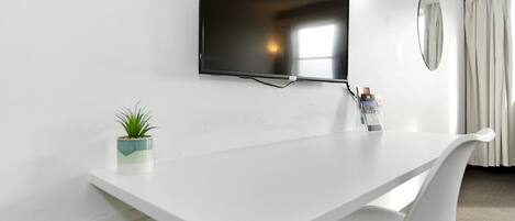 Habitación de hotel moderna con un escritorio blanco, un televisor de pantalla plana montado en la pared, una pequeña planta en maceta y un espejo redondo.