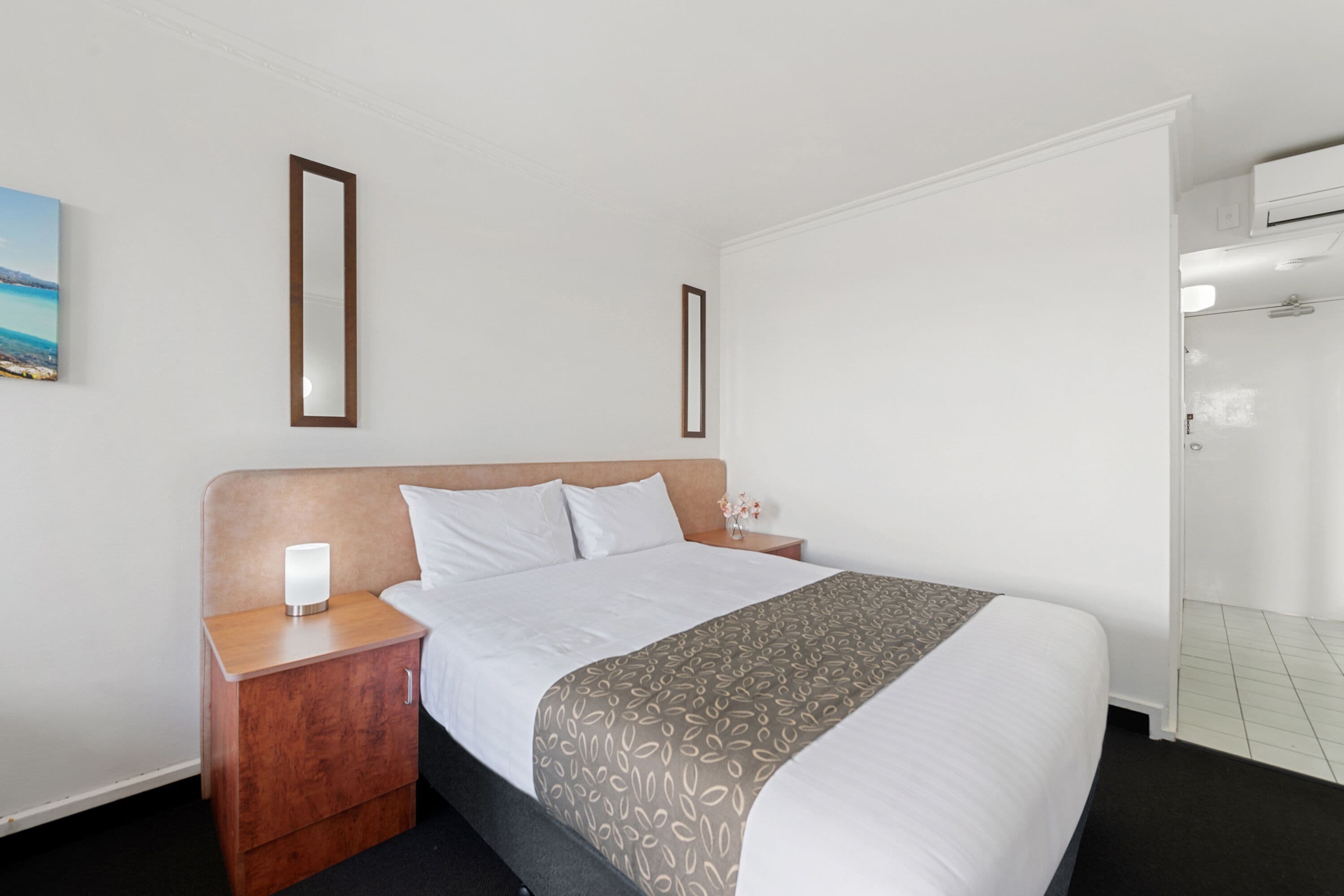 Habitación de hotel con cama, mesita de noche con lámpara, aire acondicionado instalado en la pared y un cuadro enmarcado.