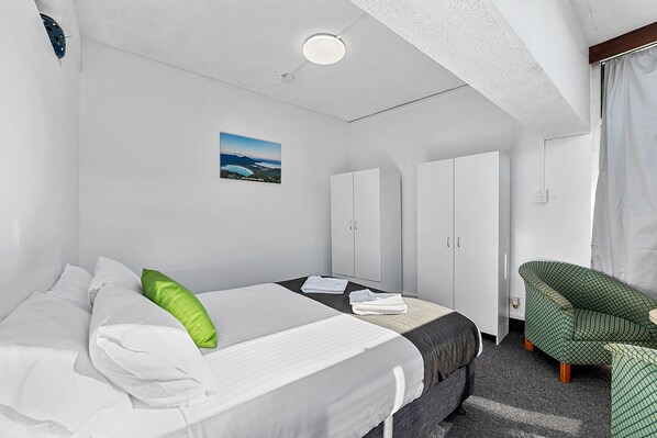 A hotel room with a bed, a green chair, a wardrobe, and a painting on the wall.