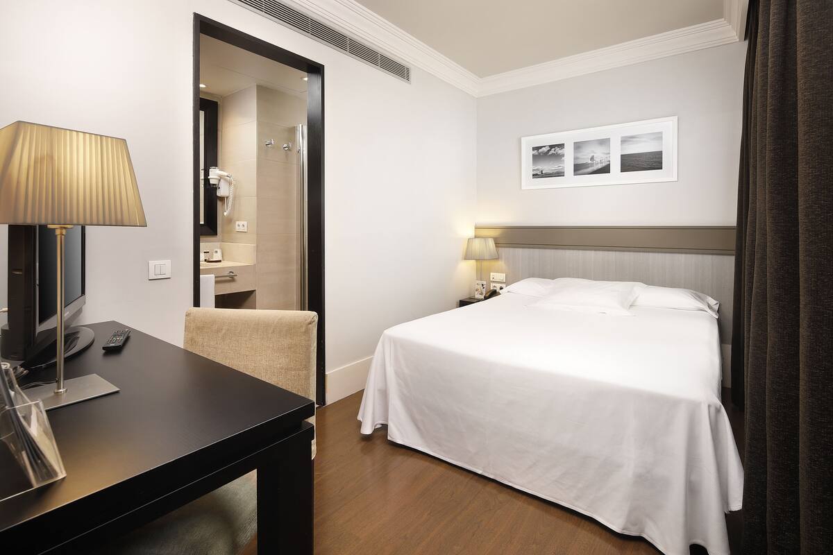 A hotel room with a bed, a desk with a lamp, a chair, a TV, and a bathroom visible through an open door.