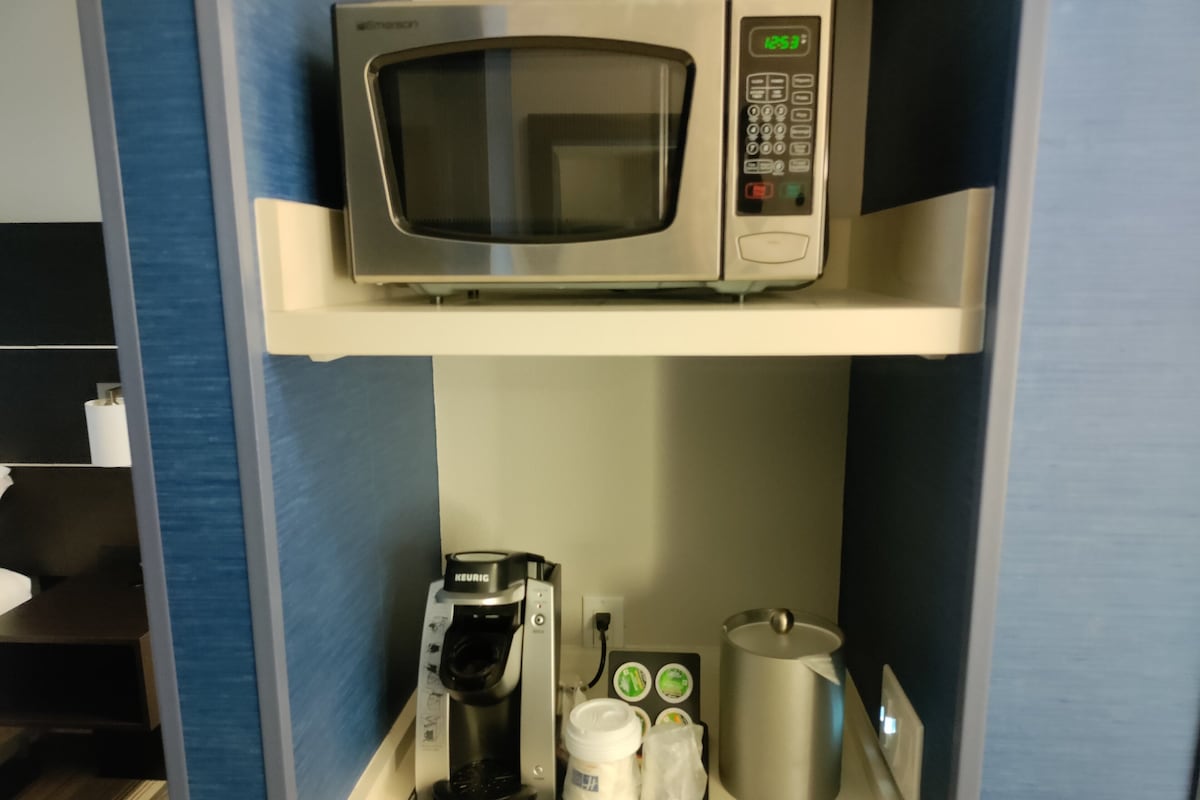 Eine Küchenzeile mit einer Mikrowelle, einer Kaffeemaschine und Kaffeekapseln auf der Arbeitsplatte.