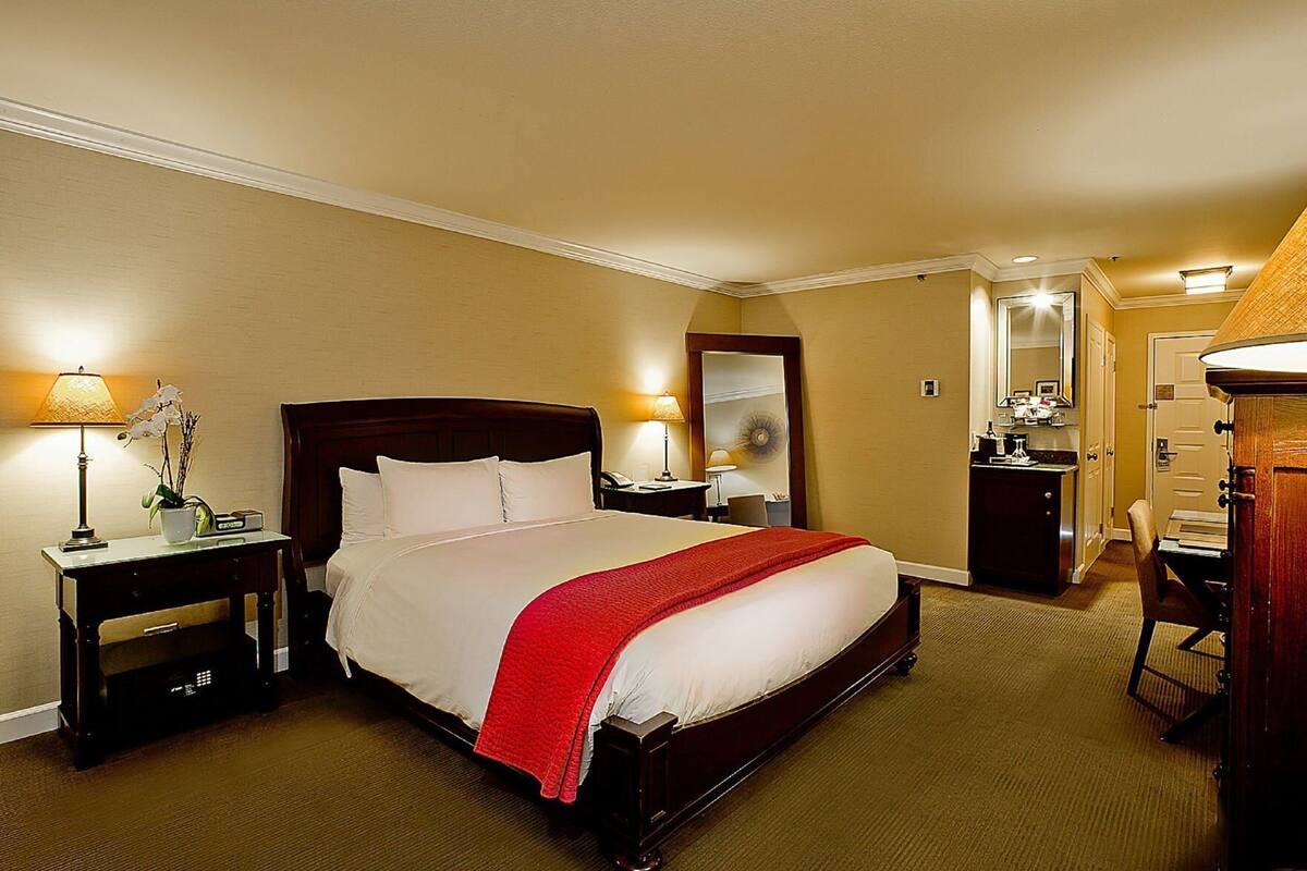 A hotel room with a large bed, bedside tables, a desk, a chair, a mirror, and a door.