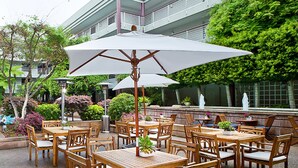 An outdoor patio with wooden tables and chairs, surrounded by lush greenery and a building in the background. - Cupertino Hotel (Cupertino)