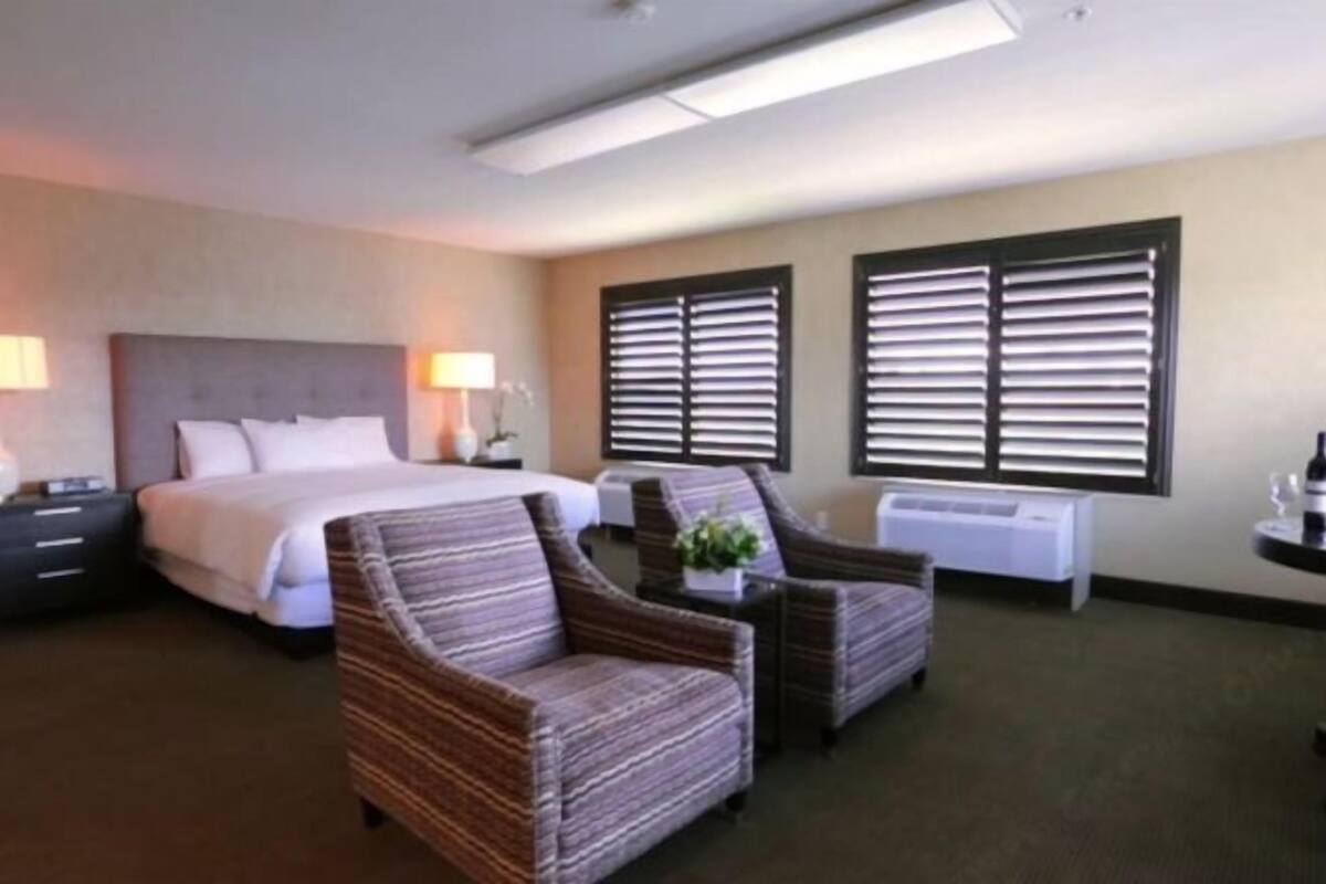 A hotel room with a bed, two armchairs, a side table, a lamp, and a window with blinds.