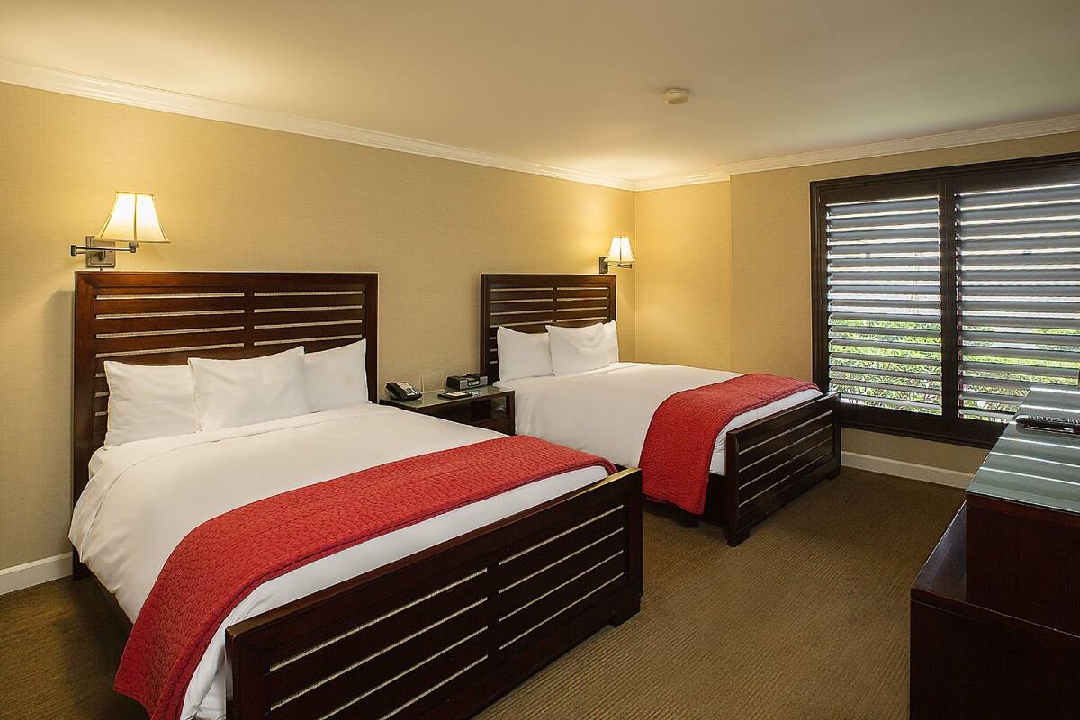 A hotel room with two beds, each with a wooden headboard, white bedding, and a red throw blanket. There are two bedside lamps and a window with blinds.