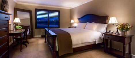 Una habitación de hotel con una cama grande, dos mesitas de noche con lámparas, un escritorio con una silla, un aparador y una ventana con persianas.