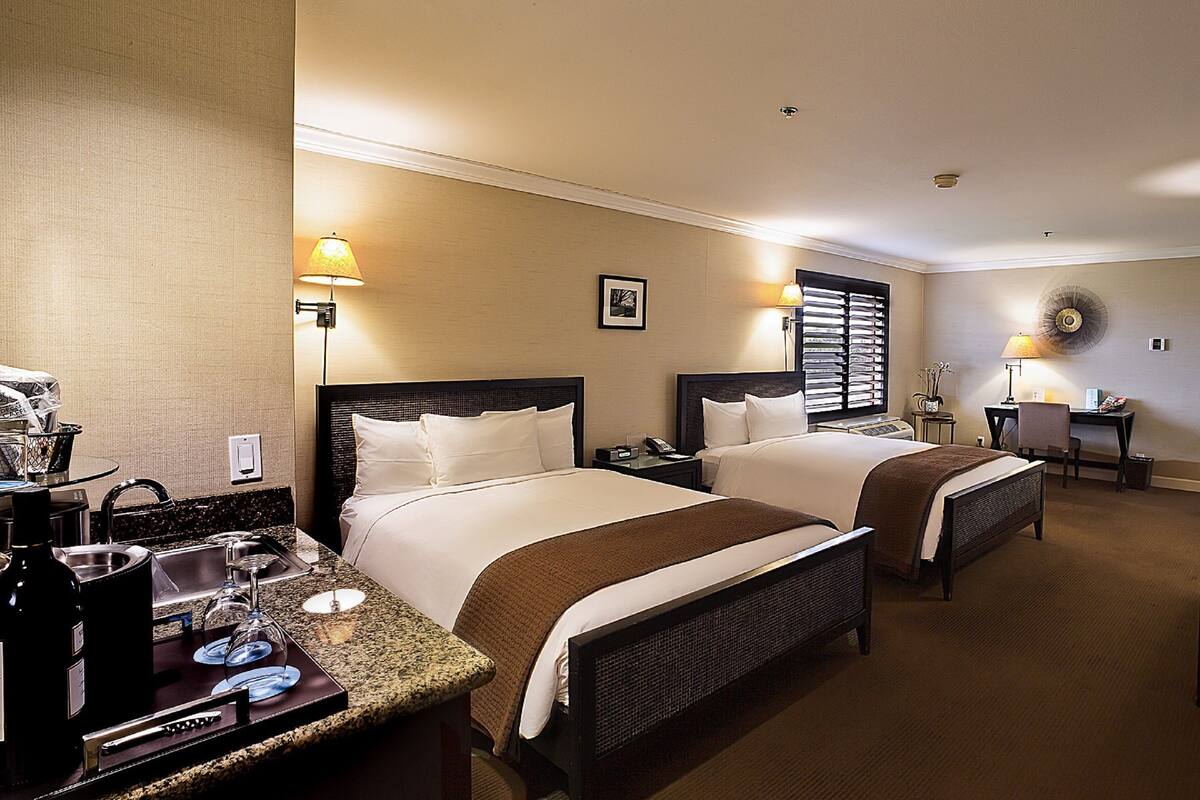 A hotel room with two beds, a desk, a chair, a small table with a lamp, and a window with blinds.