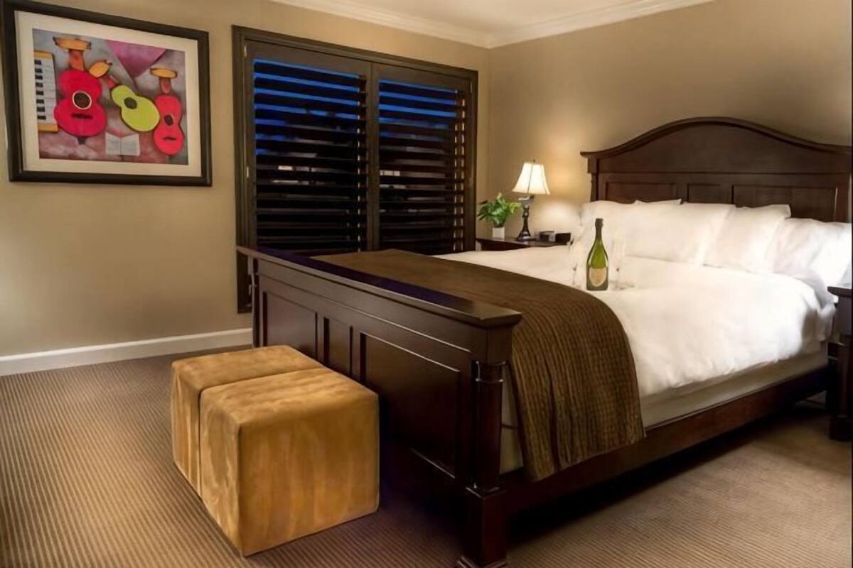 A hotel room with a bed, a nightstand, a lamp, a bottle of wine, and a painting on the wall.