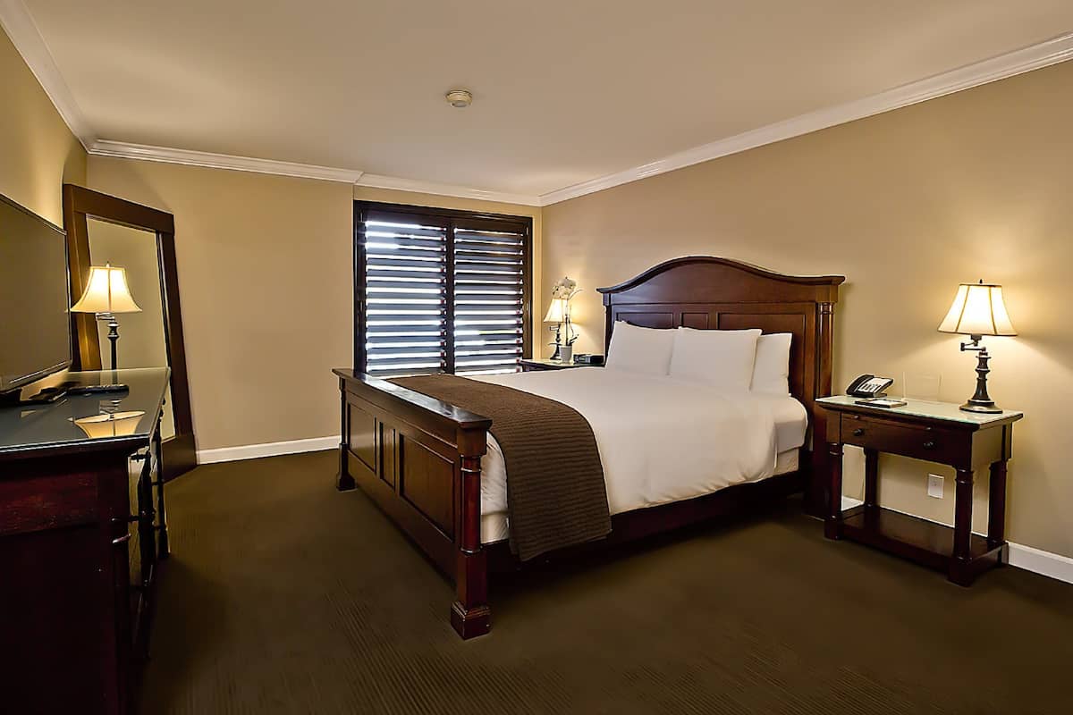 A hotel room with a large bed, bedside tables, a desk, a mirror, and a window with blinds.