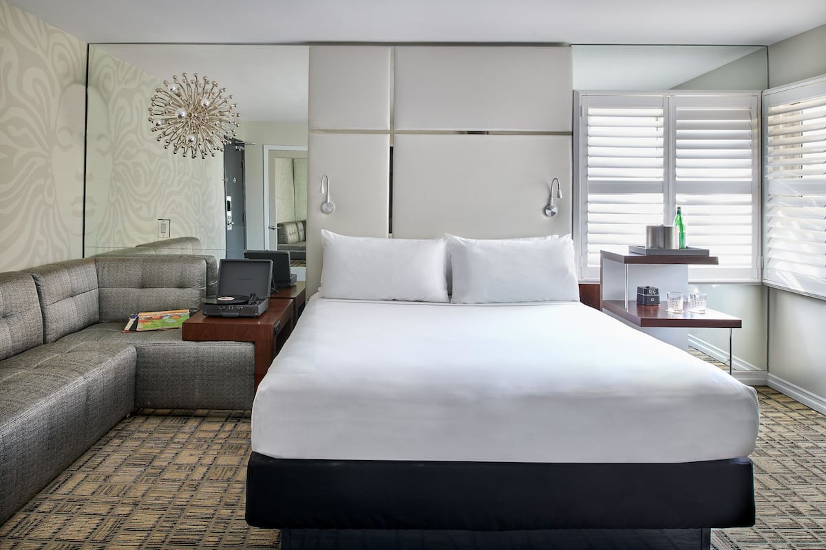 A hotel room with a large bed, a sofa, a desk with a computer, a mirror, and a window with blinds.