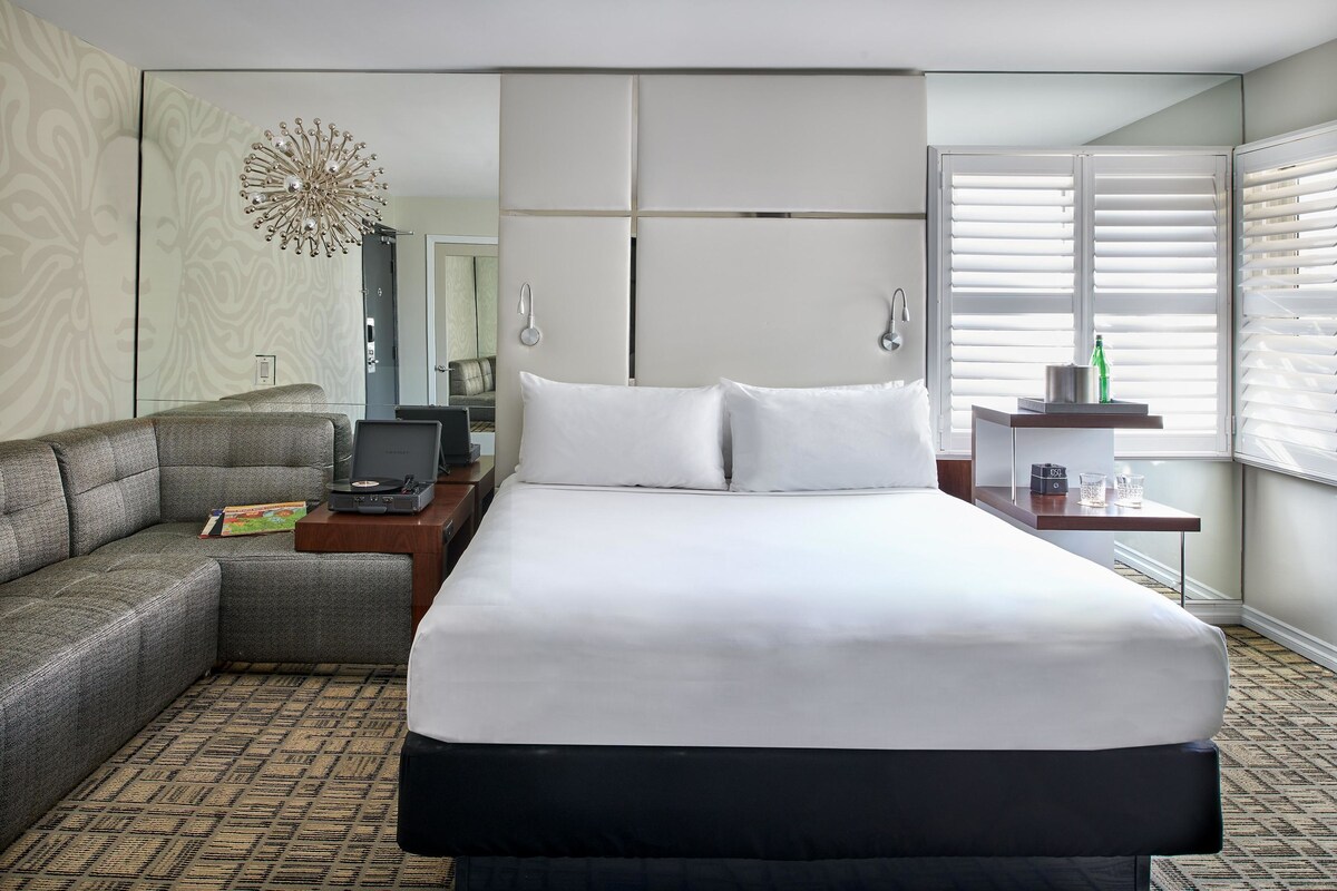 A hotel room with a large bed, a sofa, a desk with a computer, a mirror, and a window with blinds.